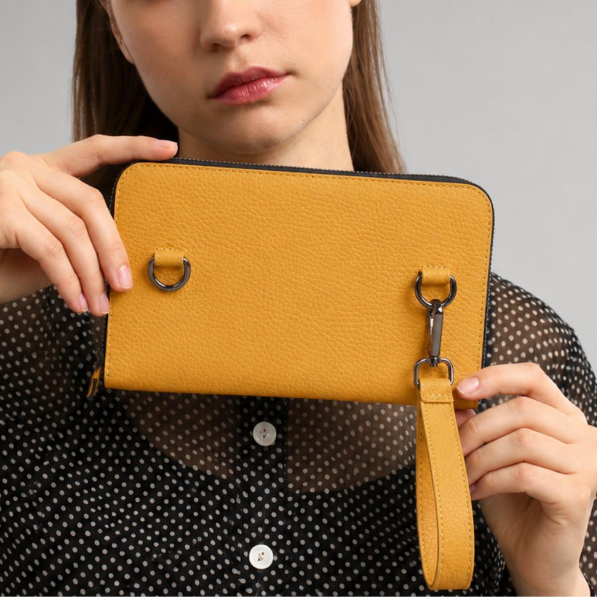 A woman holding a mustard yellow textured leather clutch with a wristlet strap and silver-tone hardware, wearing a black sheer polka-dot blouse against a neutral gray background.