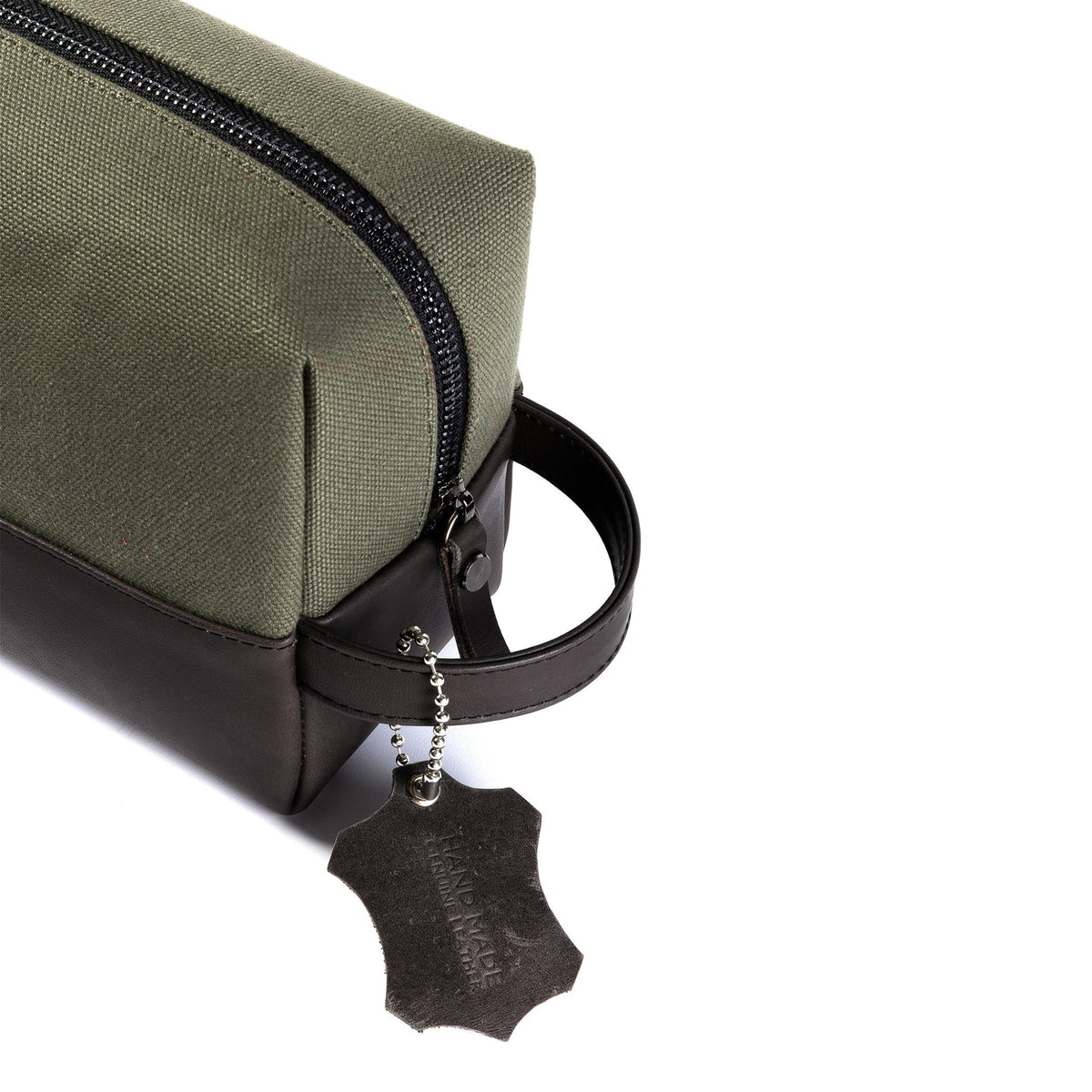 An olive green canvas zippered pouch with dark brown leather trim, a top handle, and a hanging metal tag on a plain white background.