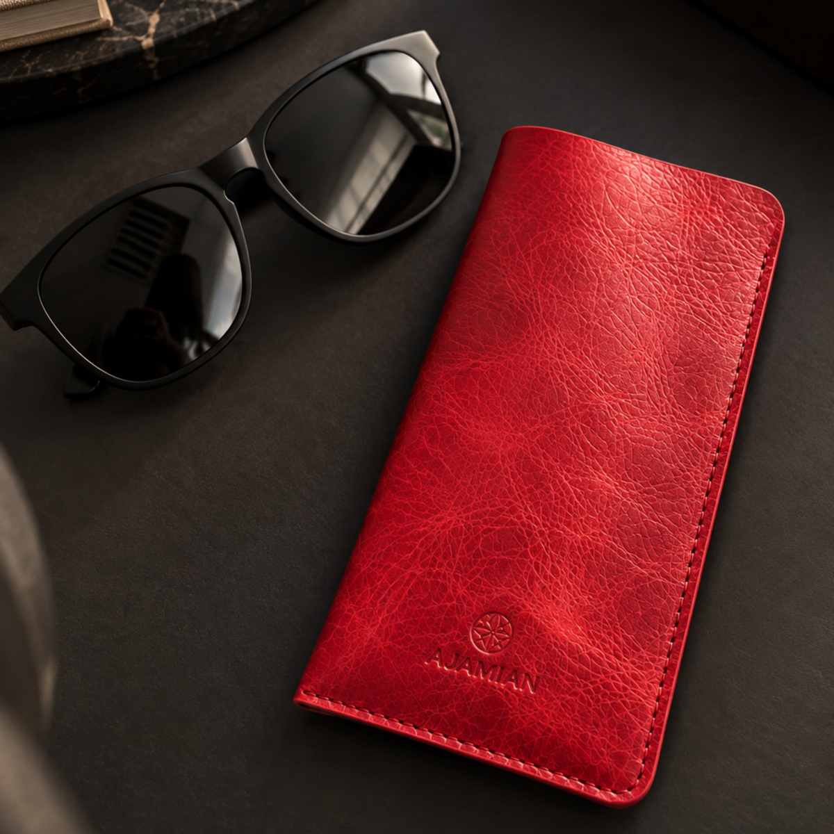 A red leather phone wallet case with a debossed logo lies on a dark textured surface next to black sunglasses. The brand name 'ALEXANDER' is embossed on the lower front of the case.