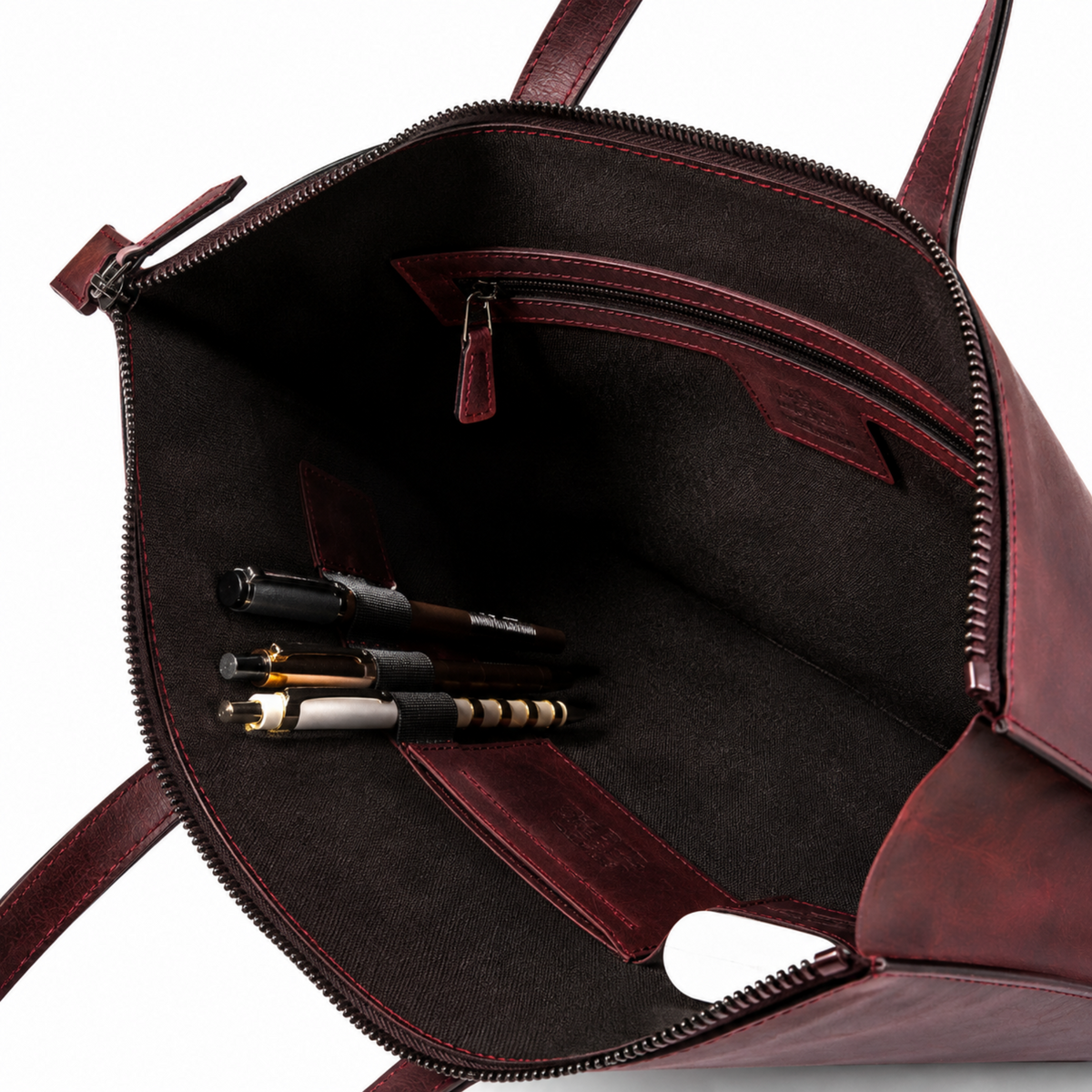 Open burgundy leather tote bag showing the interior with a dark brown fabric lining, zippered compartments, pen pockets, and several pens stored inside, photographed on a white background.