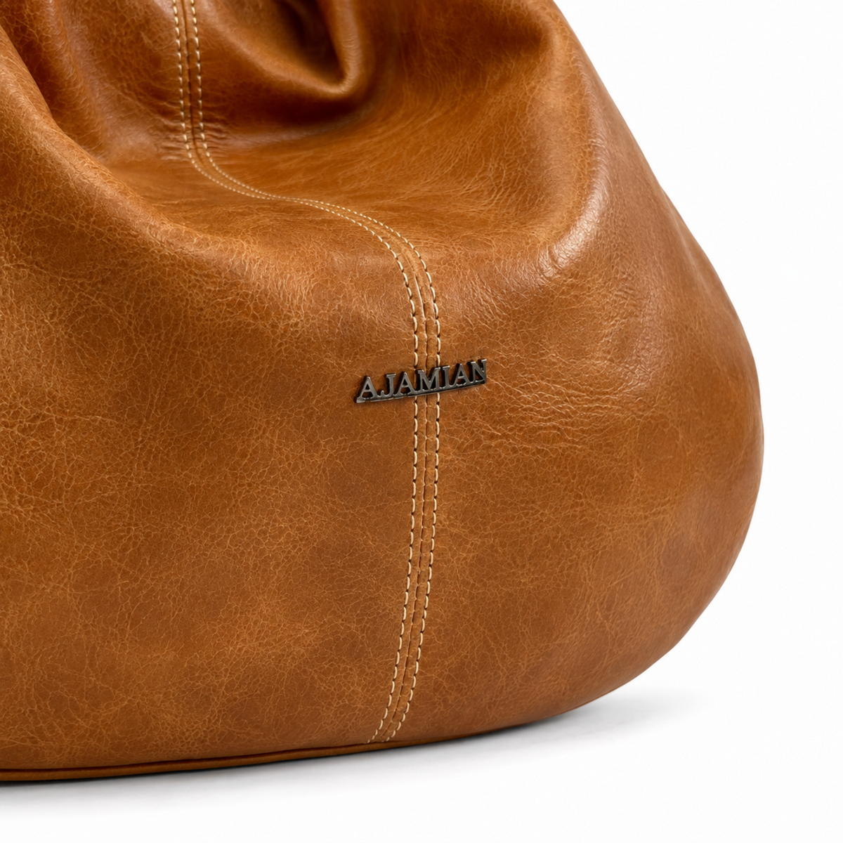 Close-up of a brown leather bag showing the texture, stitching, and a small black metal AJAMAN logo on a white background.