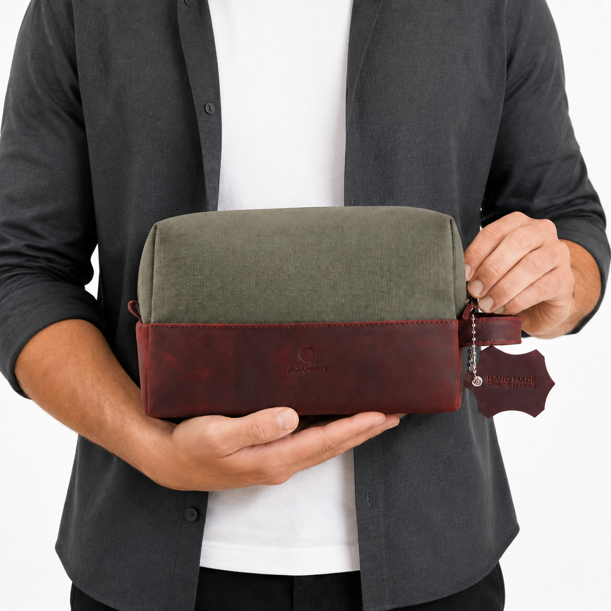 A man holds a rectangular travel pouch with an olive green canvas top and a dark reddish-brown leather bottom trim. The pouch has a matching leather zipper pull tab with the brand name 'Aurora' embossed on it.