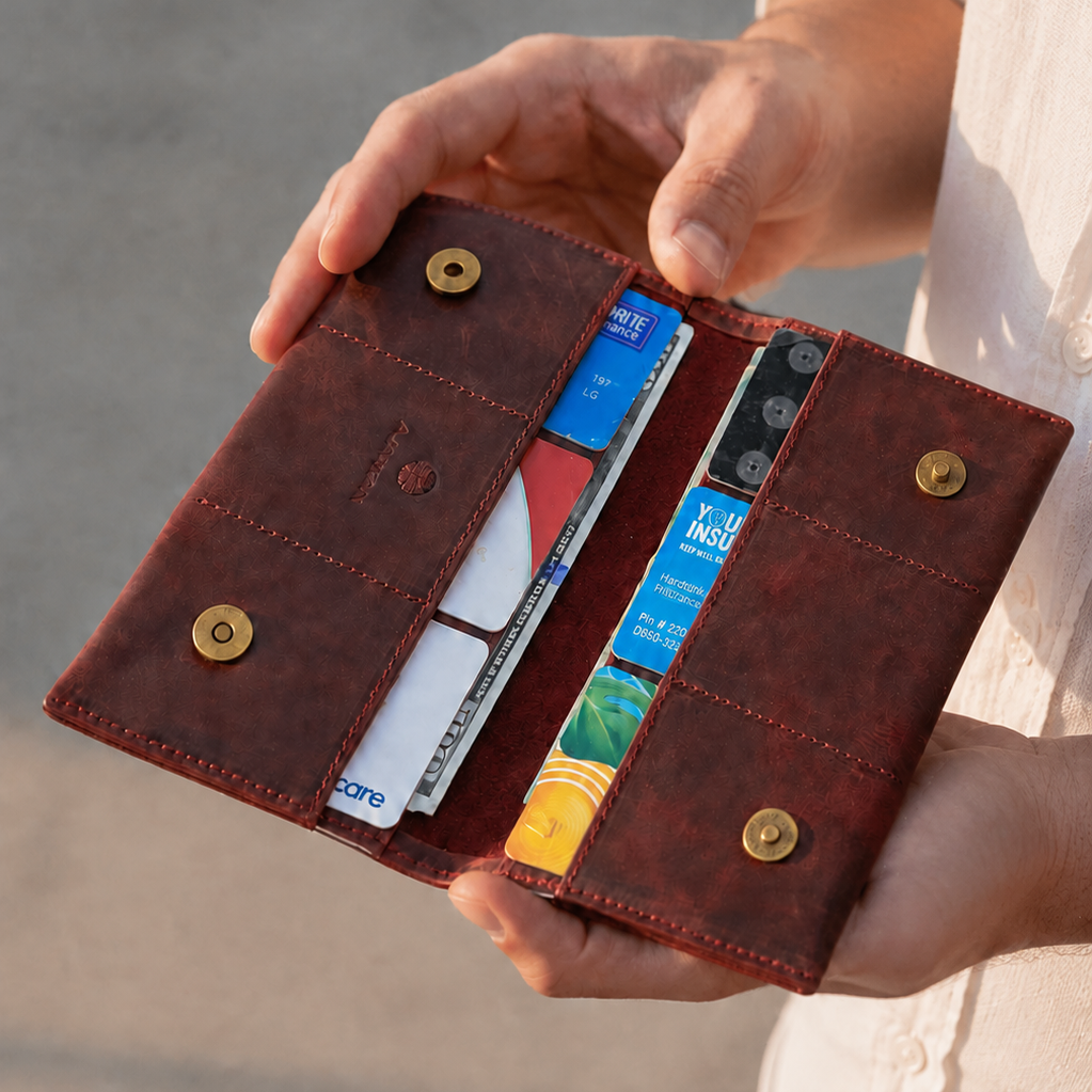 A person holds an open dark brown leather wallet with card slots and snap buttons, photographed outdoors against a blurred neutral background.
