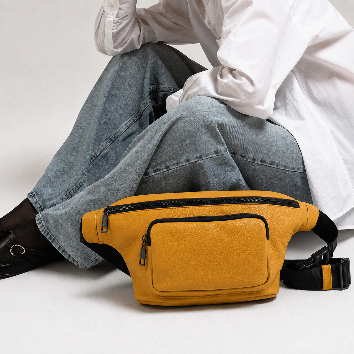 A mustard yellow textured leather fanny pack with black zippers and strap details, worn by a person in a white shirt and light blue jeans.