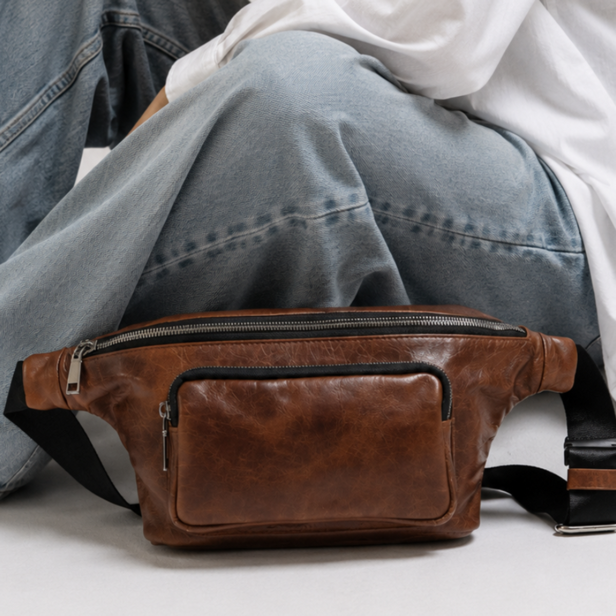 A woman wearing a white button-down shirt and light blue jeans sits cross-legged on a white floor, with a brown leather fanny pack resting on her lap.