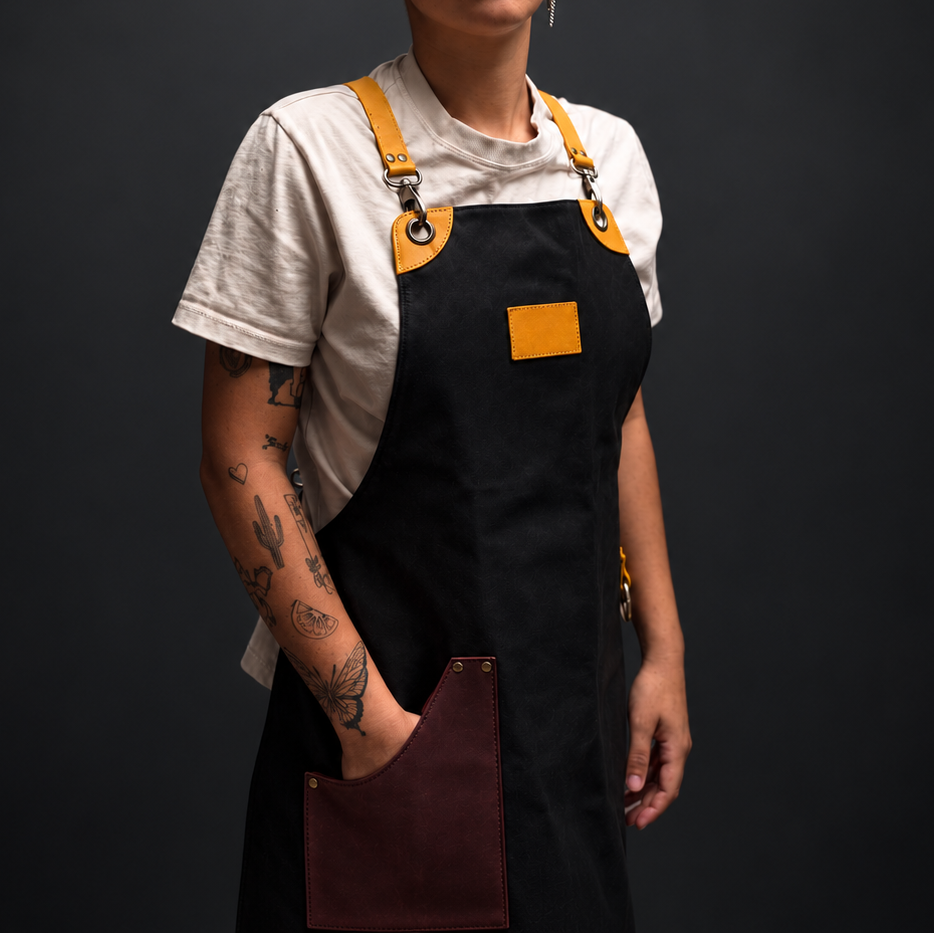 A person with short blonde hair stands against a dark studio background wearing a black apron with mustard yellow straps and a maroon front pocket over a white T-shirt.