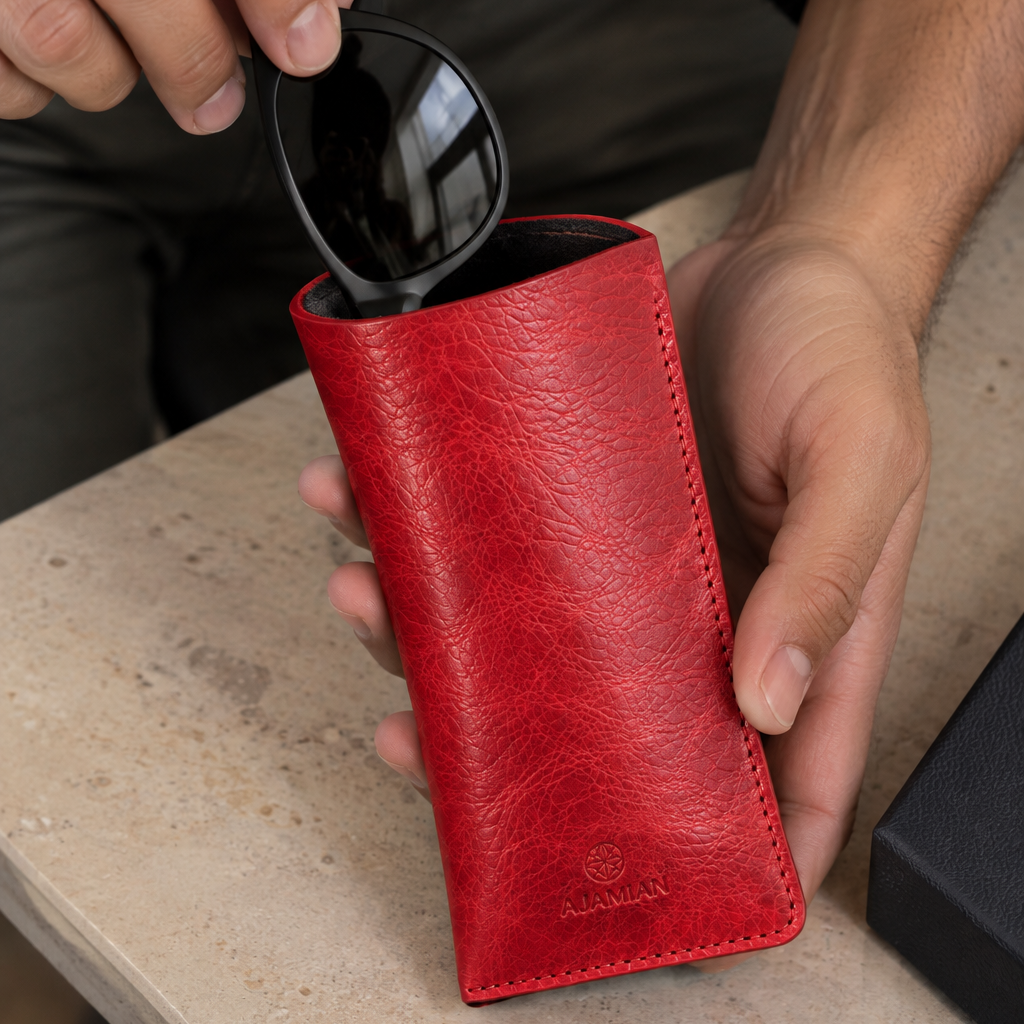 A person holds a bright red leather sunglasses case while inserting black sunglasses into it, set on a light stone tabletop.
