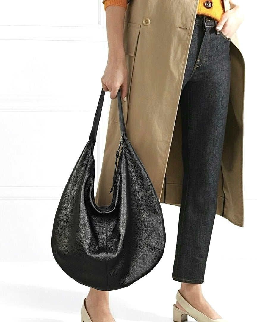 A person wearing a tan trench coat, dark gray jeans, and beige heels carries a large black pebbled leather hobo shoulder bag against a white paneled wall.