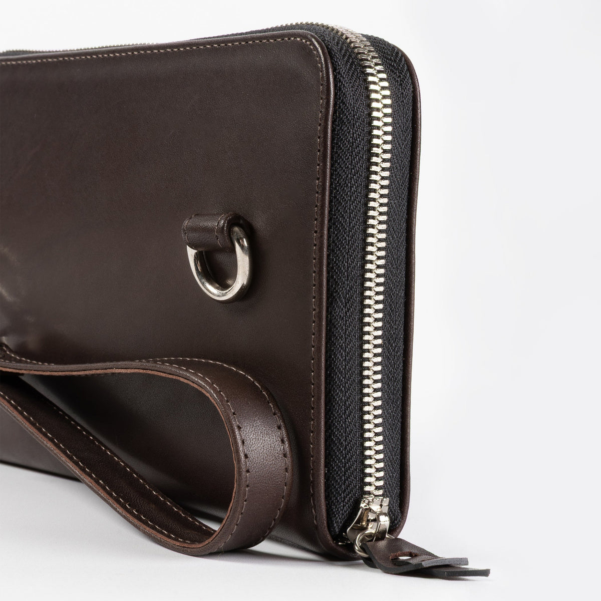 A close-up of a dark brown leather zip-around clutch or wallet with a matching wrist strap and a silver-tone metal D-ring, set against a plain light gray background.