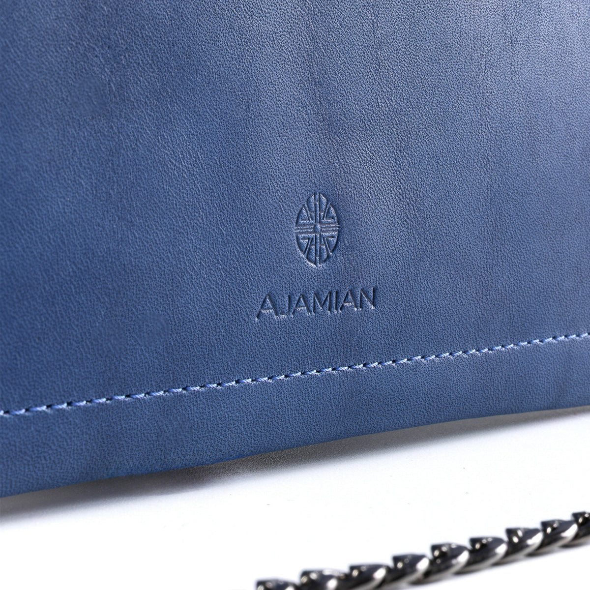 Close-up of a blue leather clutch or handbag with an embossed AJAMIAN logo and a black chain strap visible at the bottom right, set against a white background.