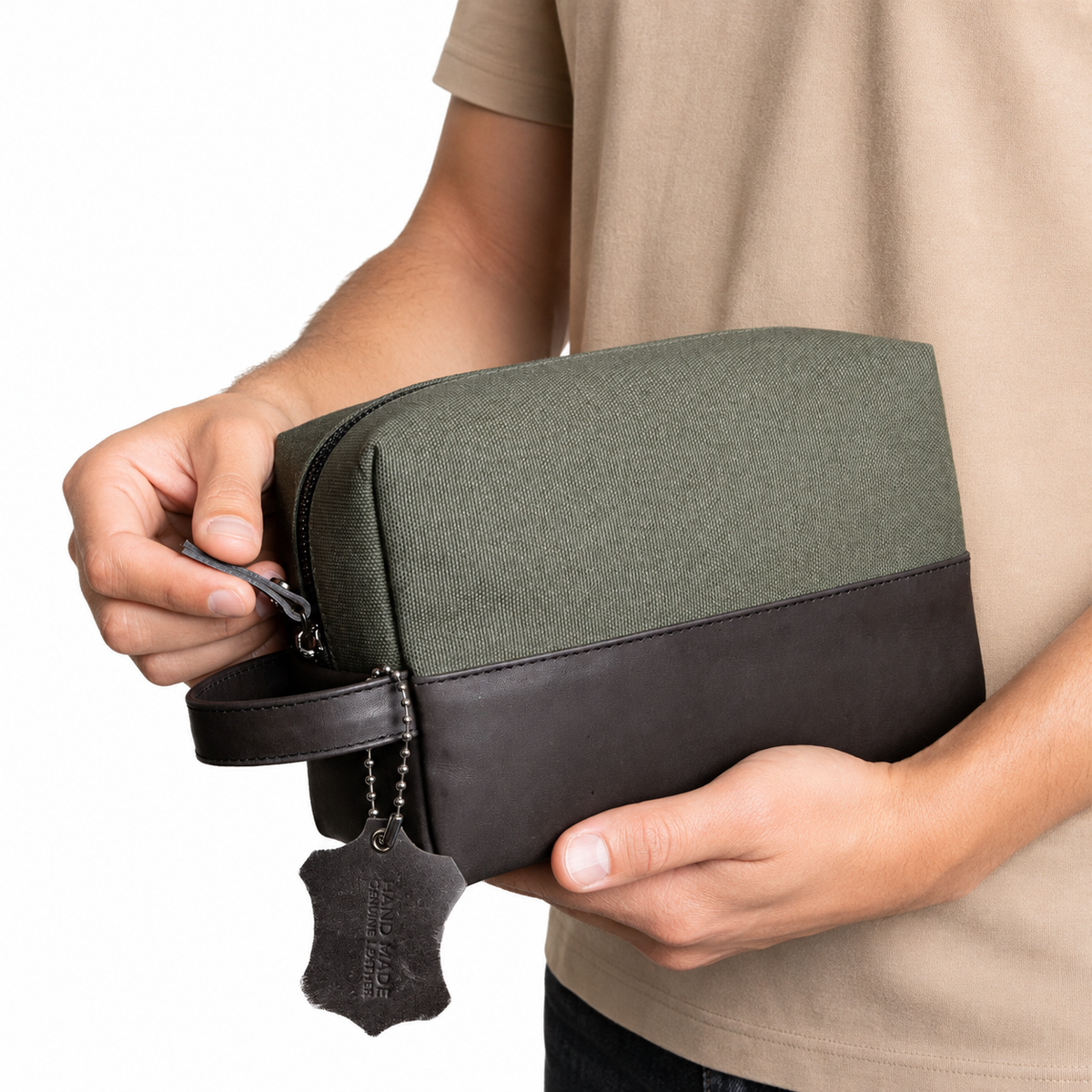A person holding a rectangular zippered pouch with an olive green canvas upper section and a dark brown leather lower section and handle. A hanging leather tag is attached, and the person is wearing a beige t-shirt against a plain white background.
