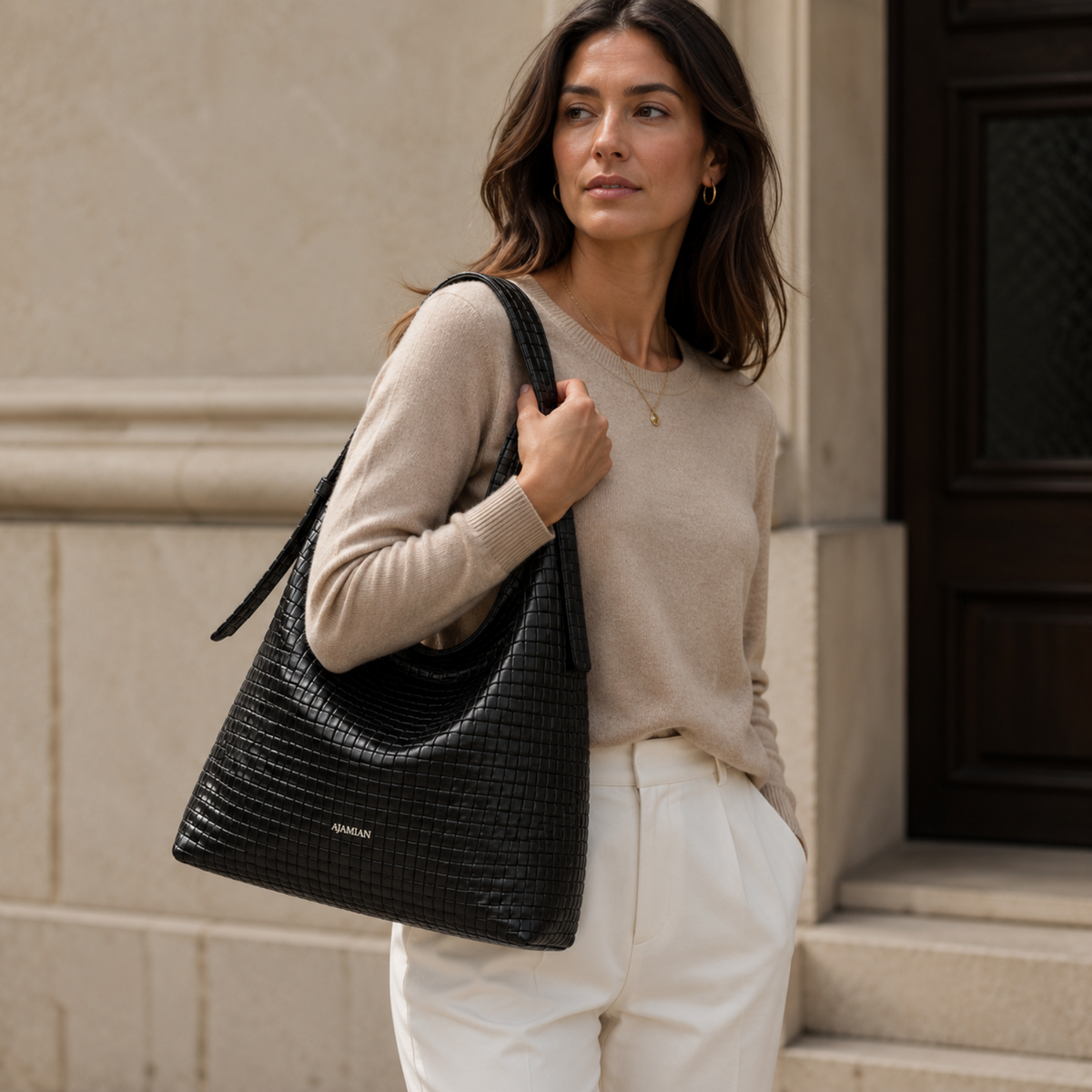 A woman wearing a beige sweater and white trousers carries a black croc-embossed shoulder bag with a small gold logo that reads 'AMIRLUN' against a beige stone building background.