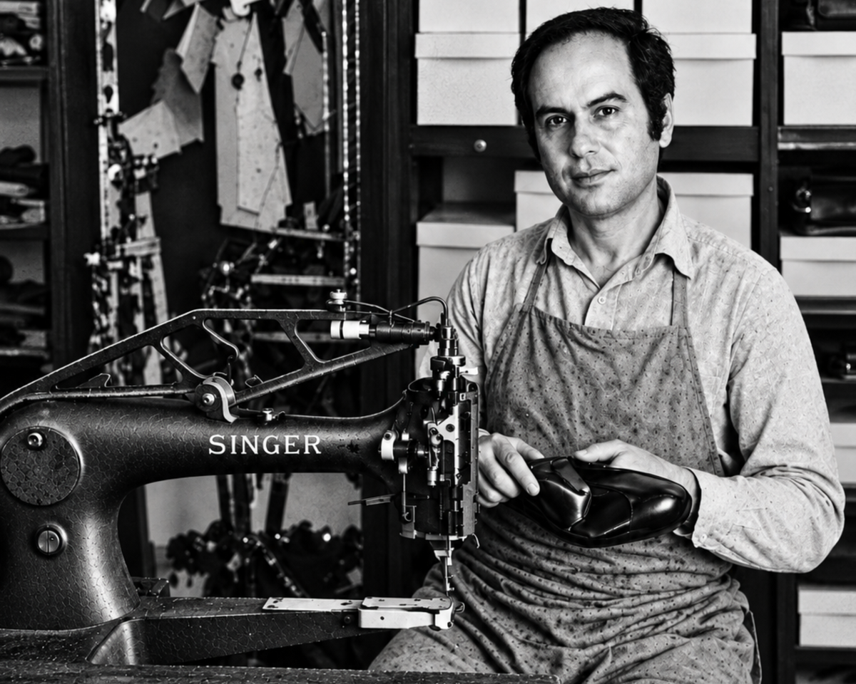 A black and white photograph of a man wearing an apron and holding a black leather shoe or boot while seated beside a vintage Singer sewing machine in a workshop filled with shelves and tools.